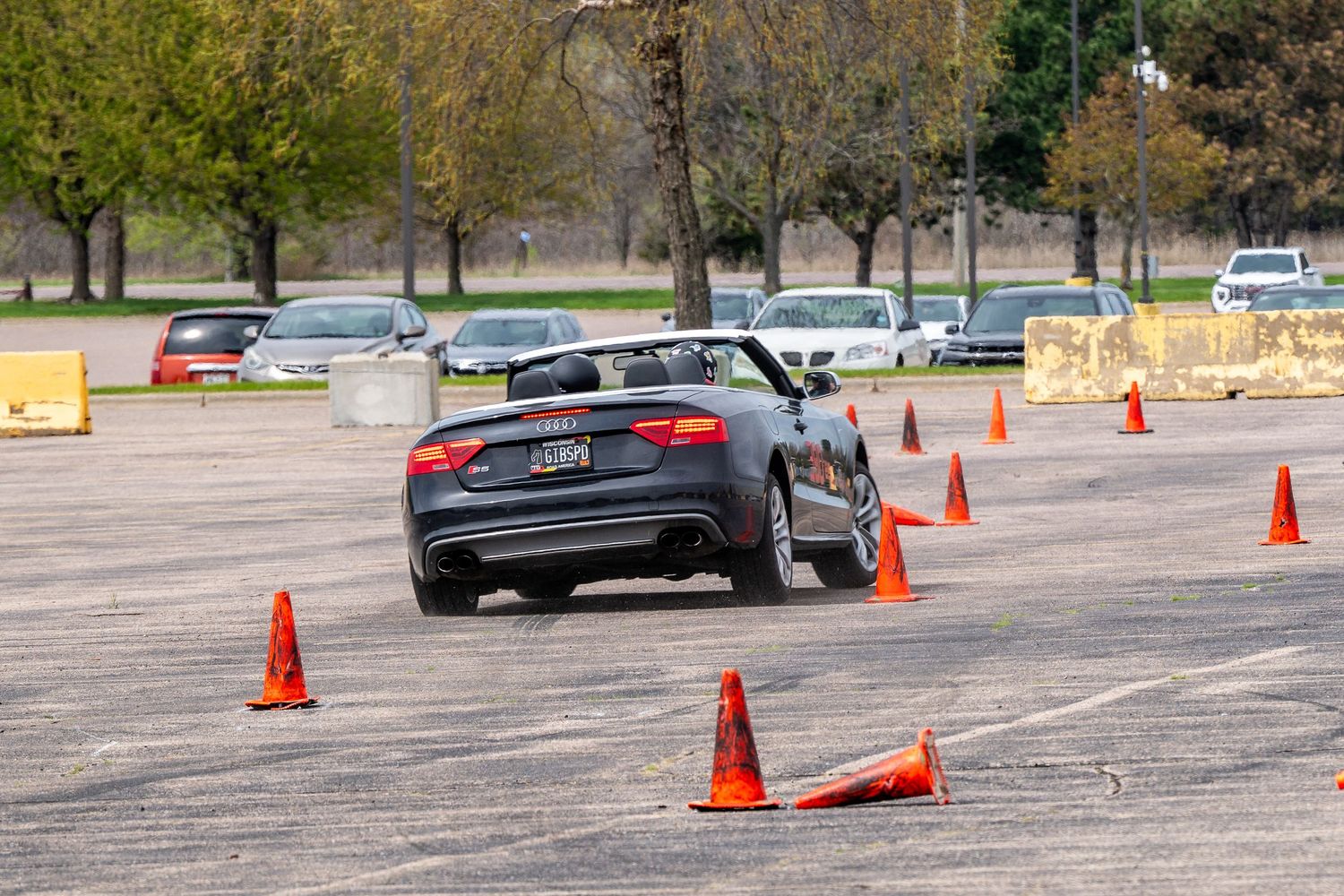 A black sports car navigates orange traffic cones on an autocross course in a parking lot during a driving event.
