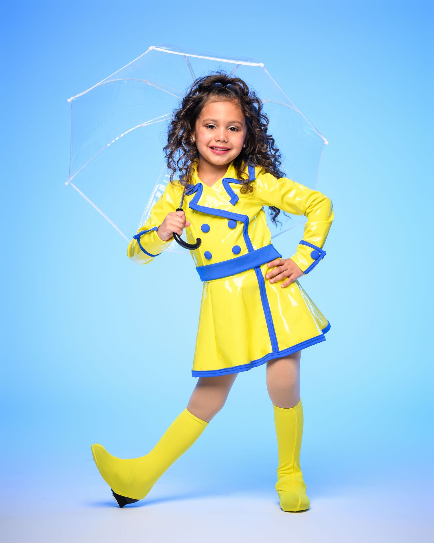 Young girl in yellow raincoat, boots, and clear umbrella poses confidently on light blue background.