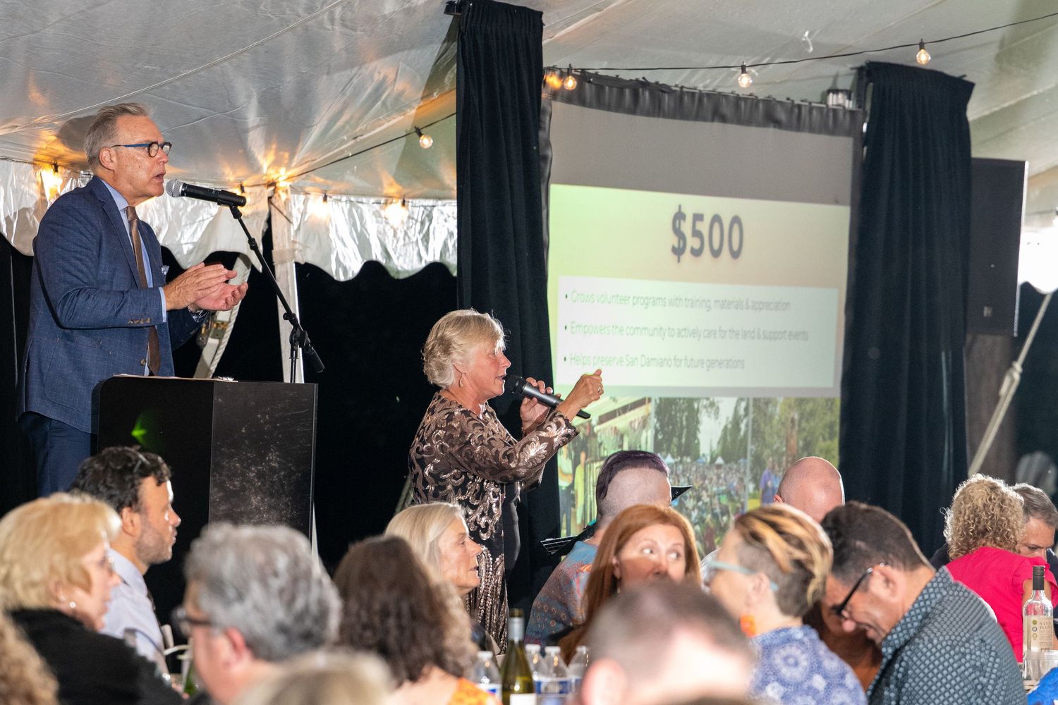 Auctioneer and bidder at a charity gala event under a tent with a $500 bid displayed on a projection screen.