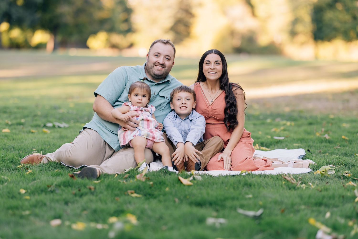 Family of four smiling on a blanket in a sunny park, parents with two young children enjoying outdoor time together.