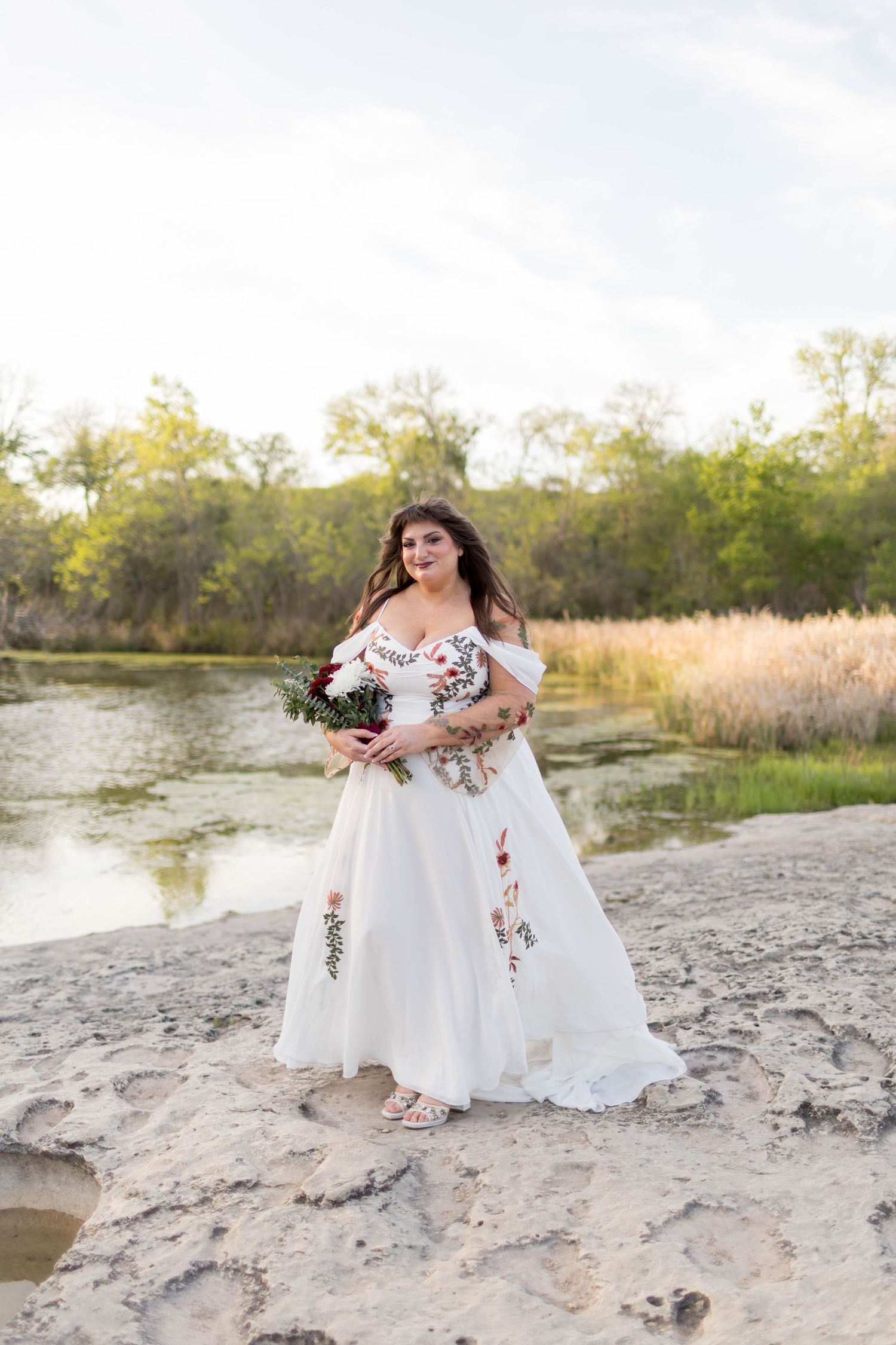 Bride in white floral embroidered off-shoulder gown walks sunlit forest path holding bouquet taken at McKinney Falls State Park.