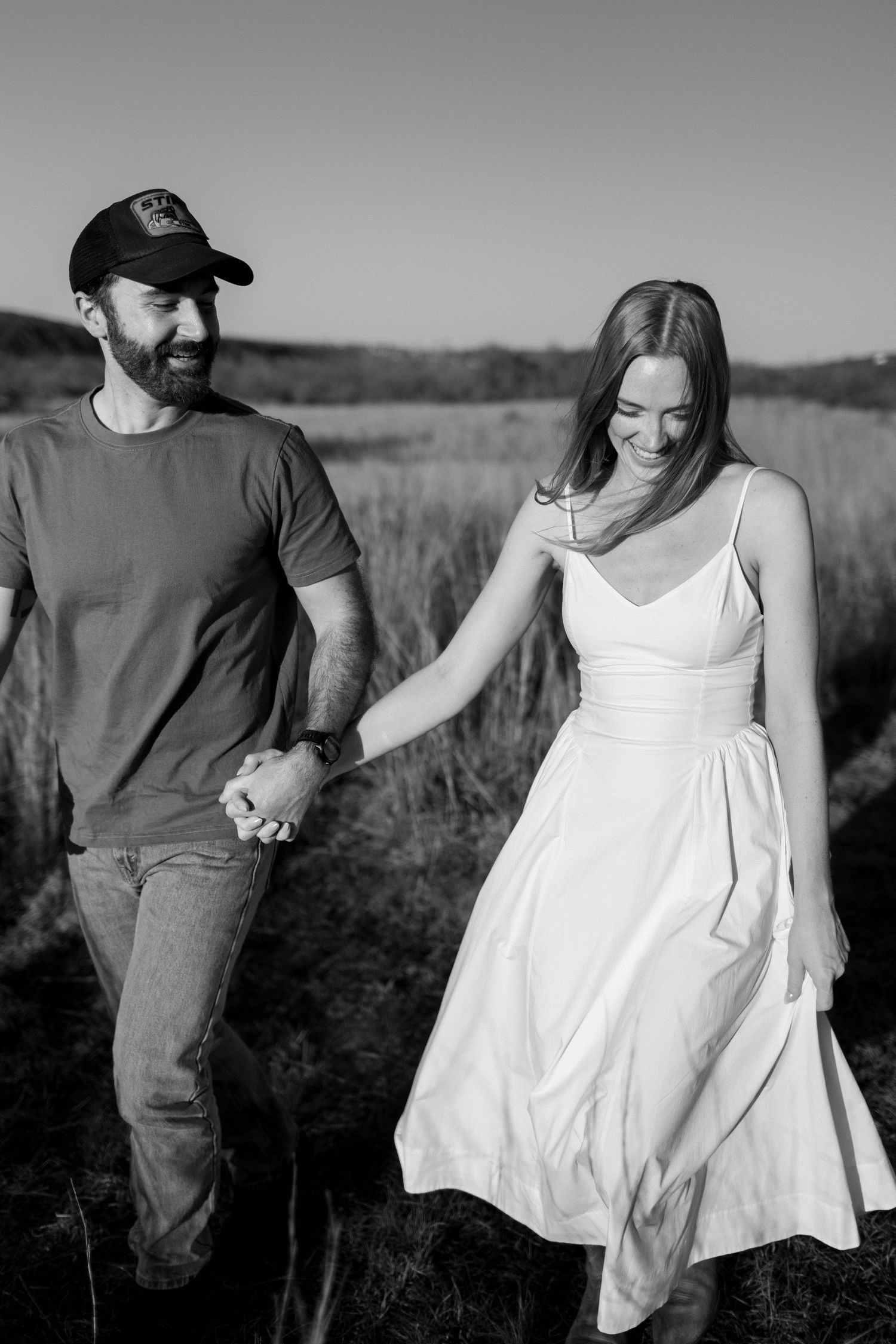 A smiling couple holds hands walking through a golden field, woman in white dress, man in gray tee and cap.