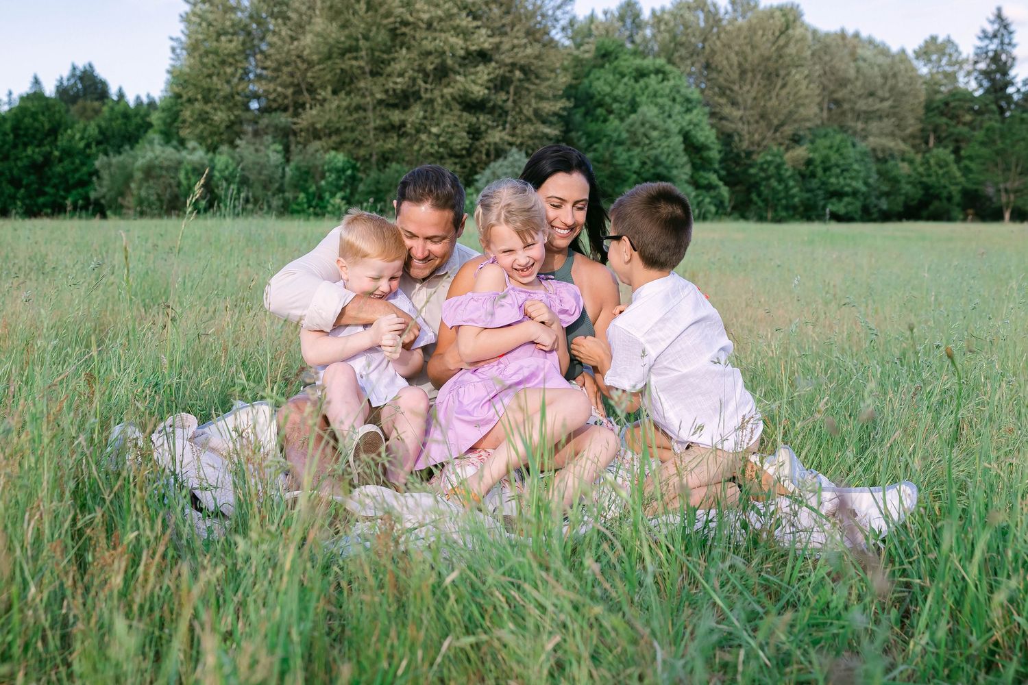 Happy family sitting together in a green meadow, parents and three children laughing outdoors on a sunny day.