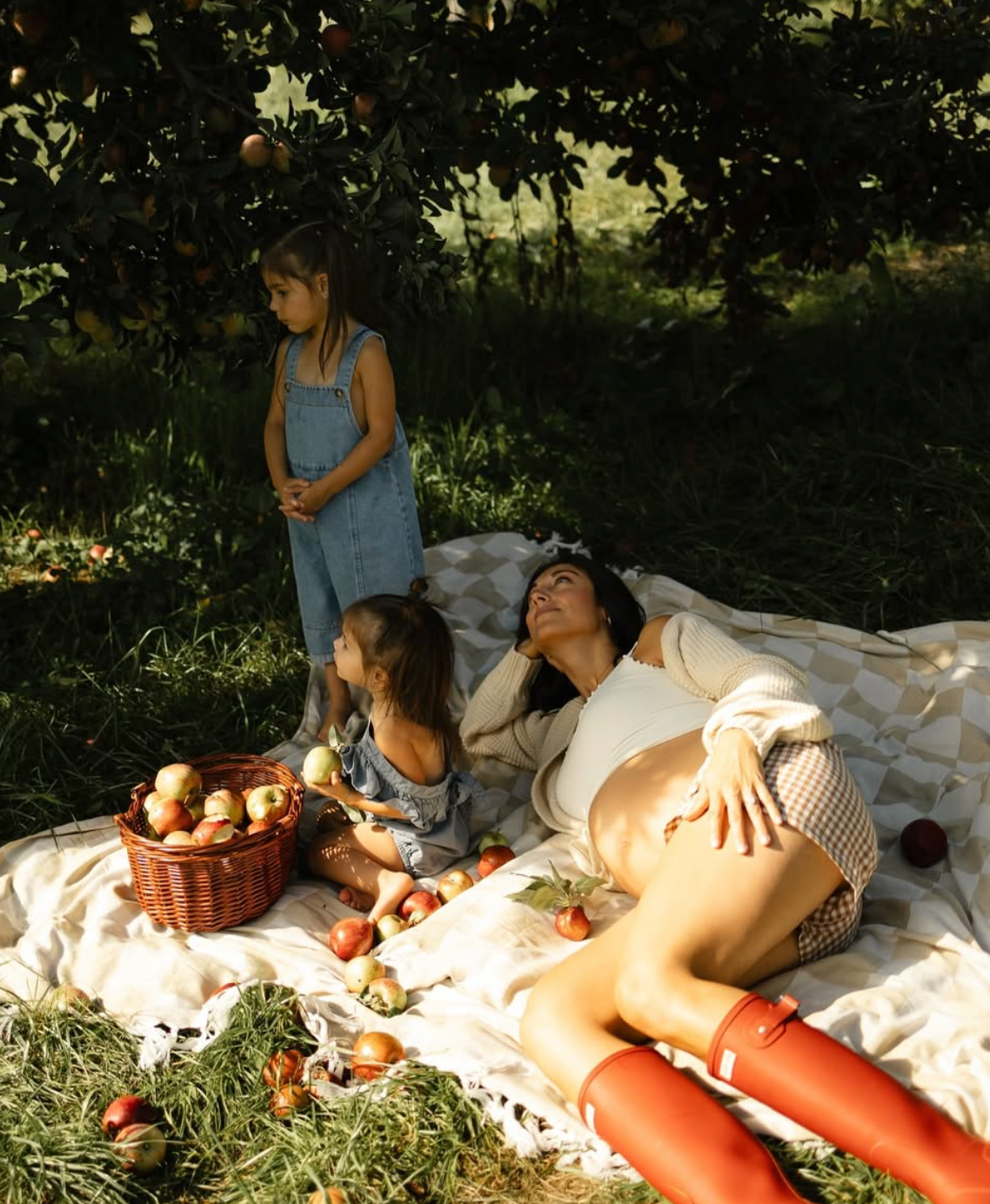 Family picnic in an apple orchard with a woman reclining on a blanket and two young girls surrounded by fresh apples.