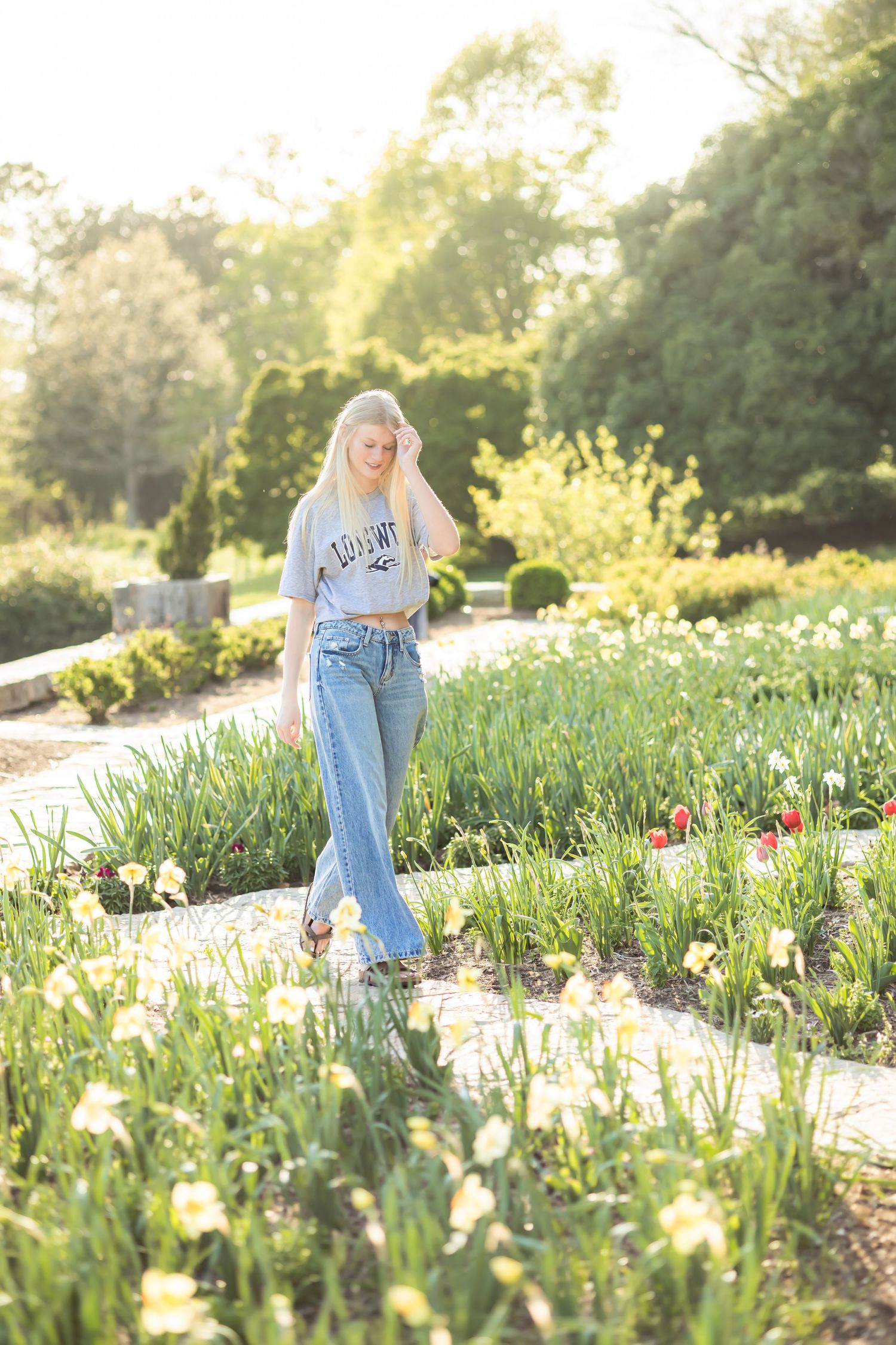Blonde woman in jeans and gray tee walks among spring flowers in a sunlit garden path.