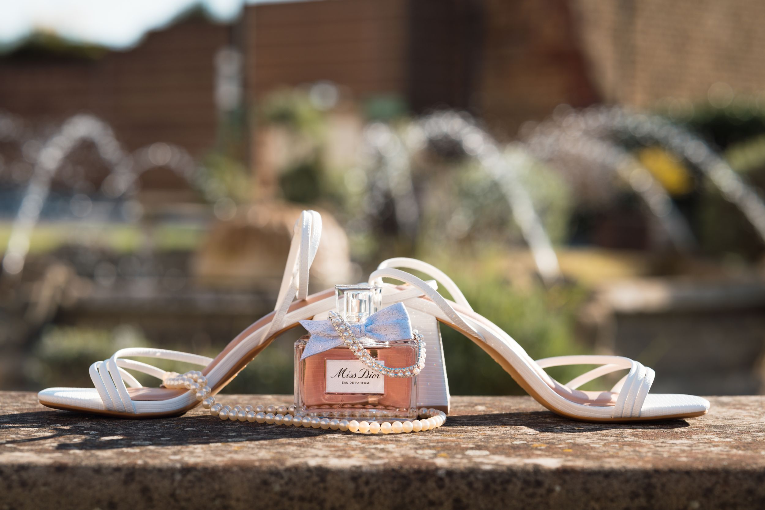 Crystal-embellished bridal heels beside a bottle of 909 eau de parfum and pearl drop earrings on a wooden surface.