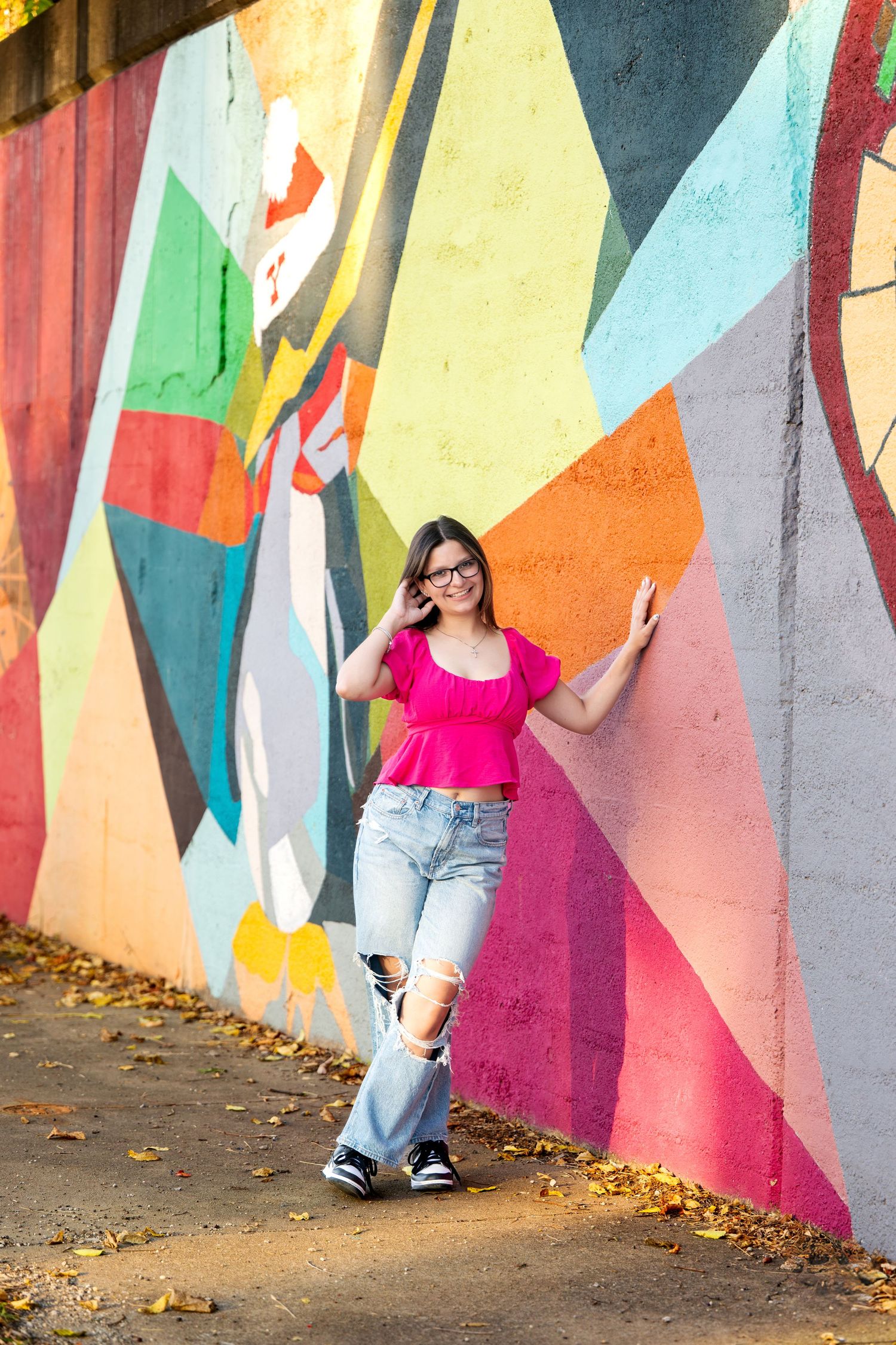 Young woman in pink crop top and ripped jeans posing against colorful graffiti wall outdoors.