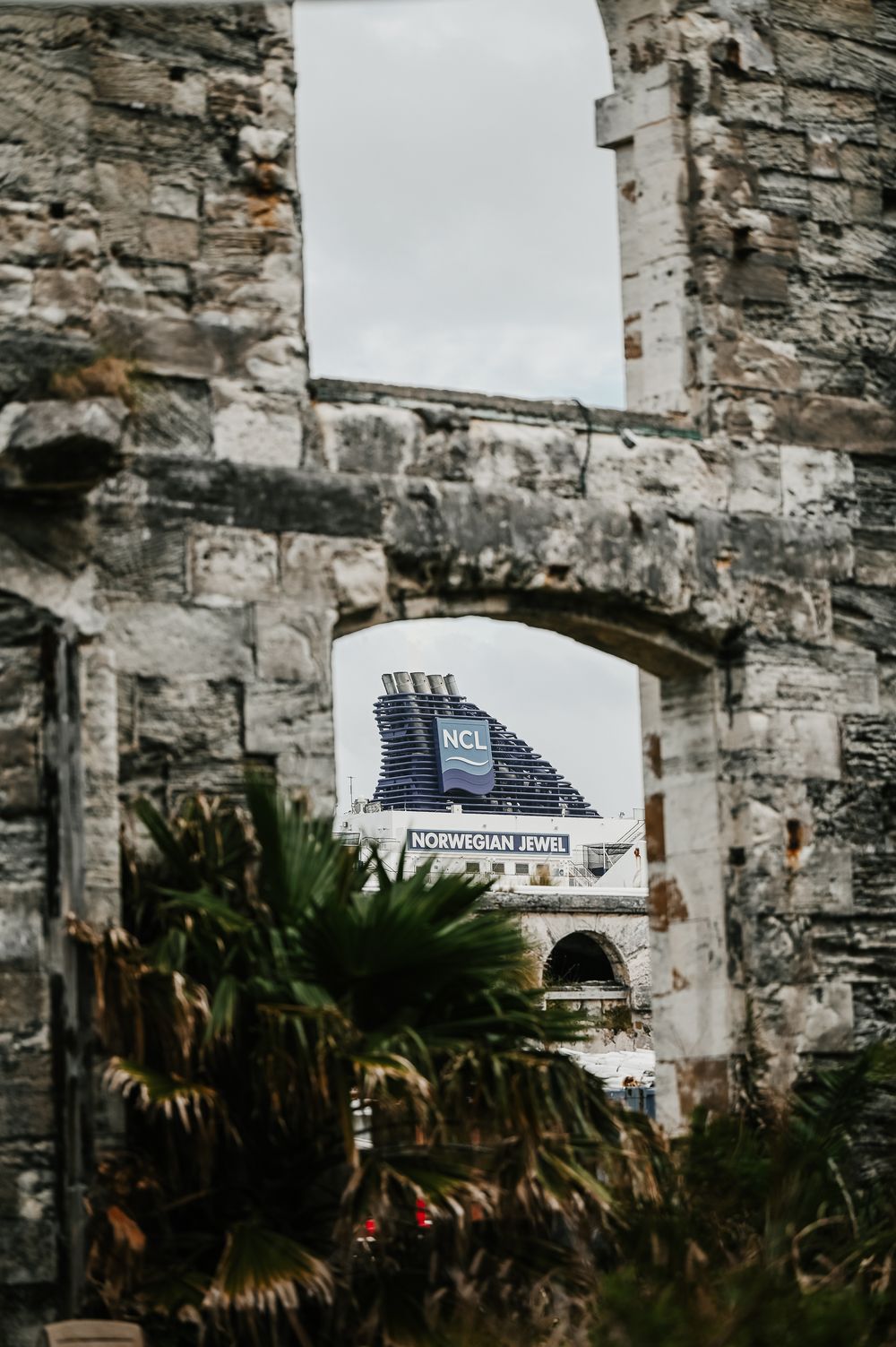 Norwegian Jewel cruise ship funnel viewed through ancient stone ruins archway with palm trees.