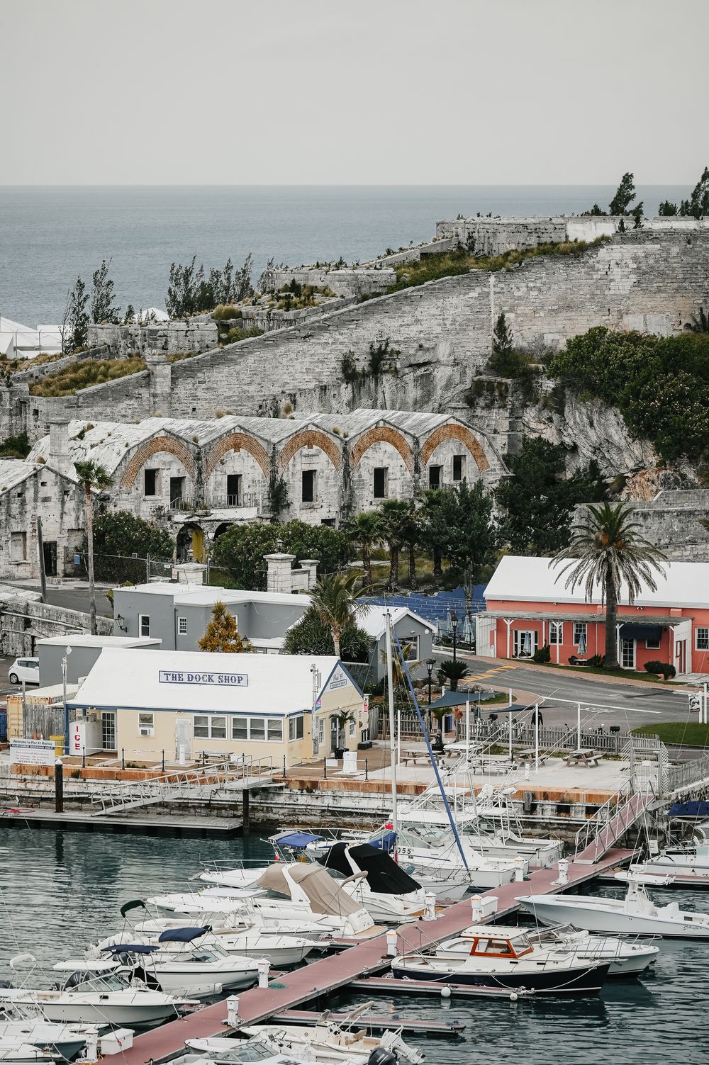 Scenic Bermuda harbor with white boats docked at marina, colorful buildings, and historic stone arches on hillside.