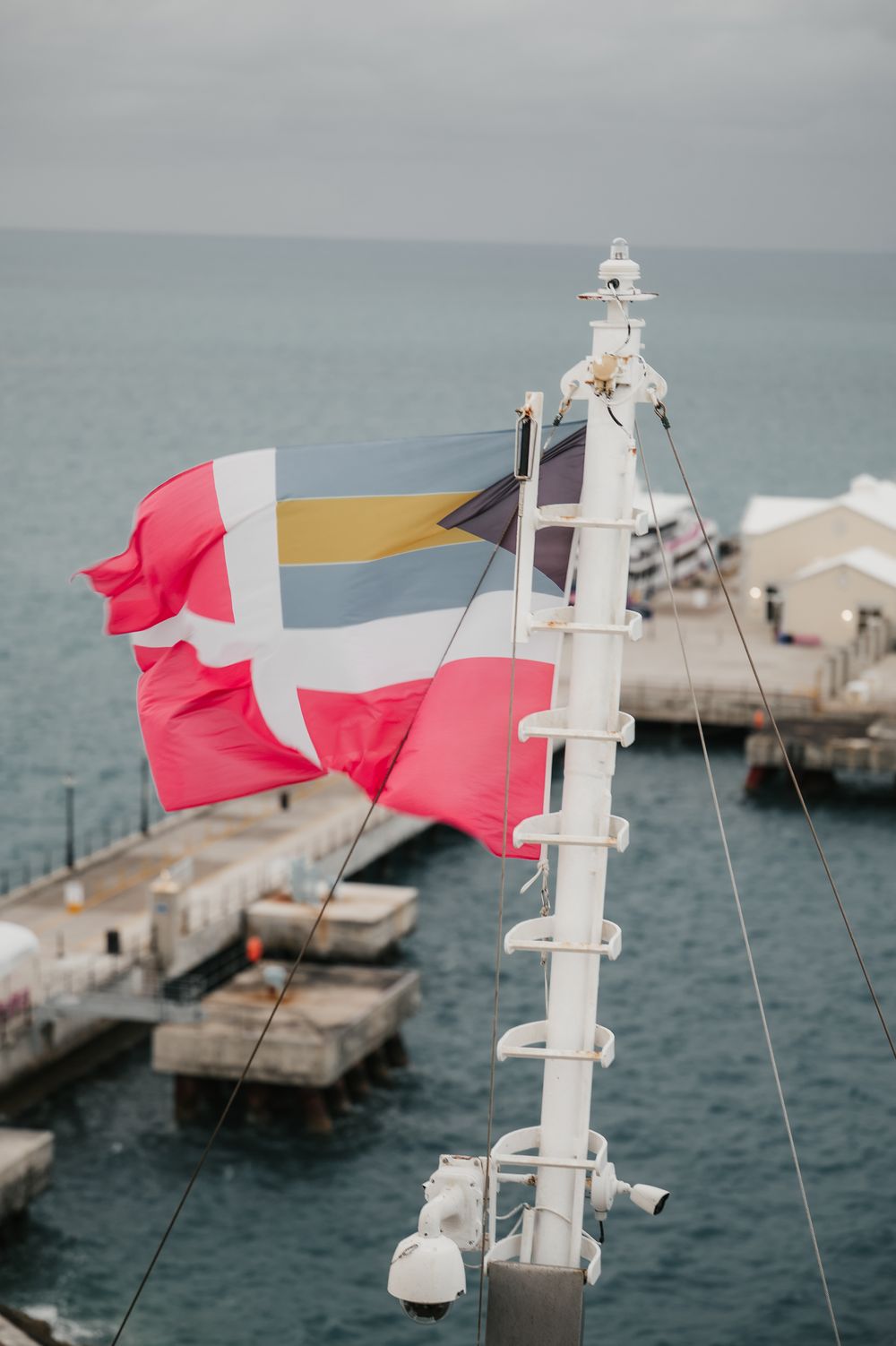 Colorful flags waving on a ship mast with a harbor and overcast sky in the background.