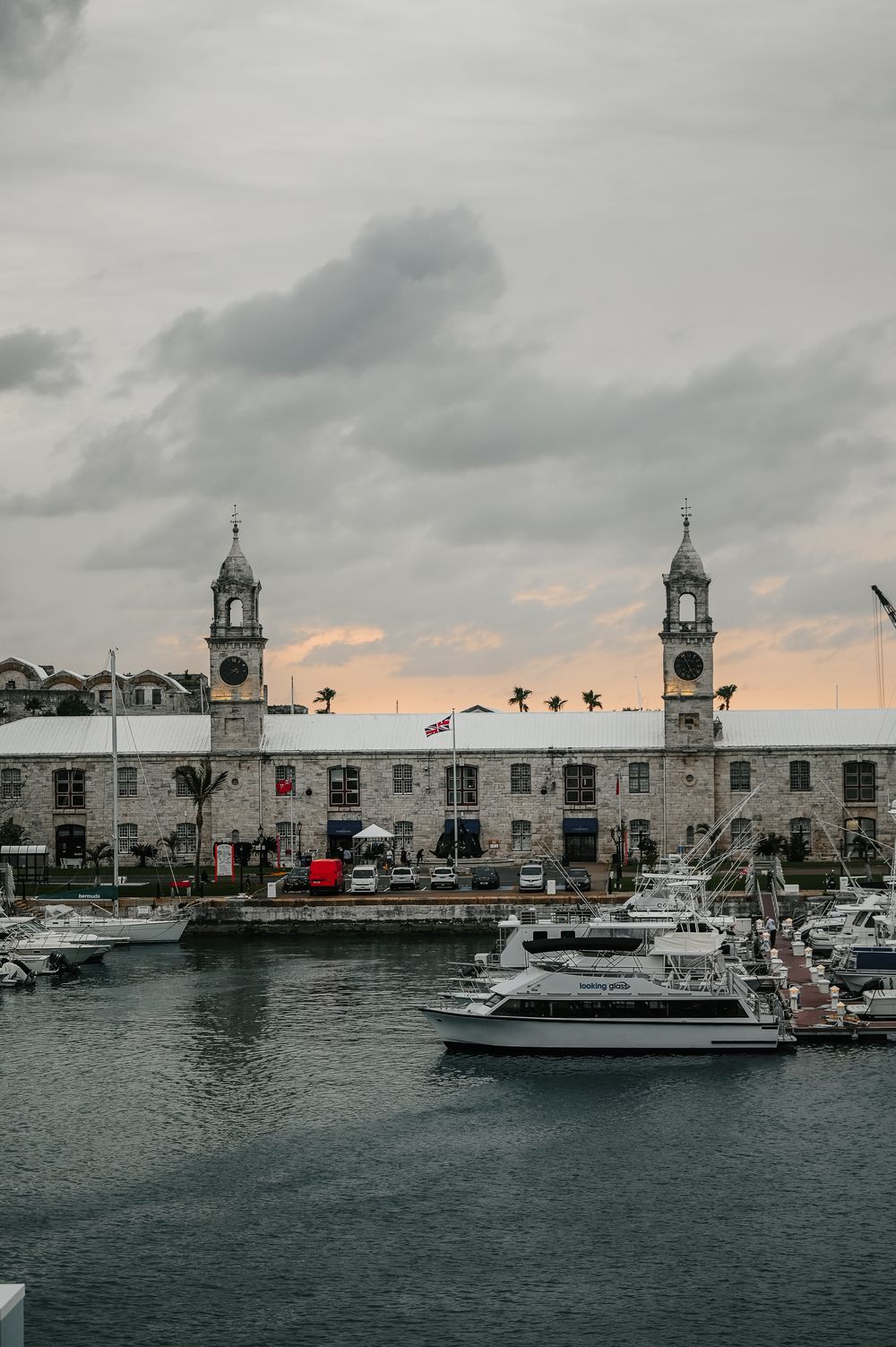Historic Bermuda marina with twin clock towers, yachts docked at waterfront, palm trees, and dramatic cloudy sunset sky.