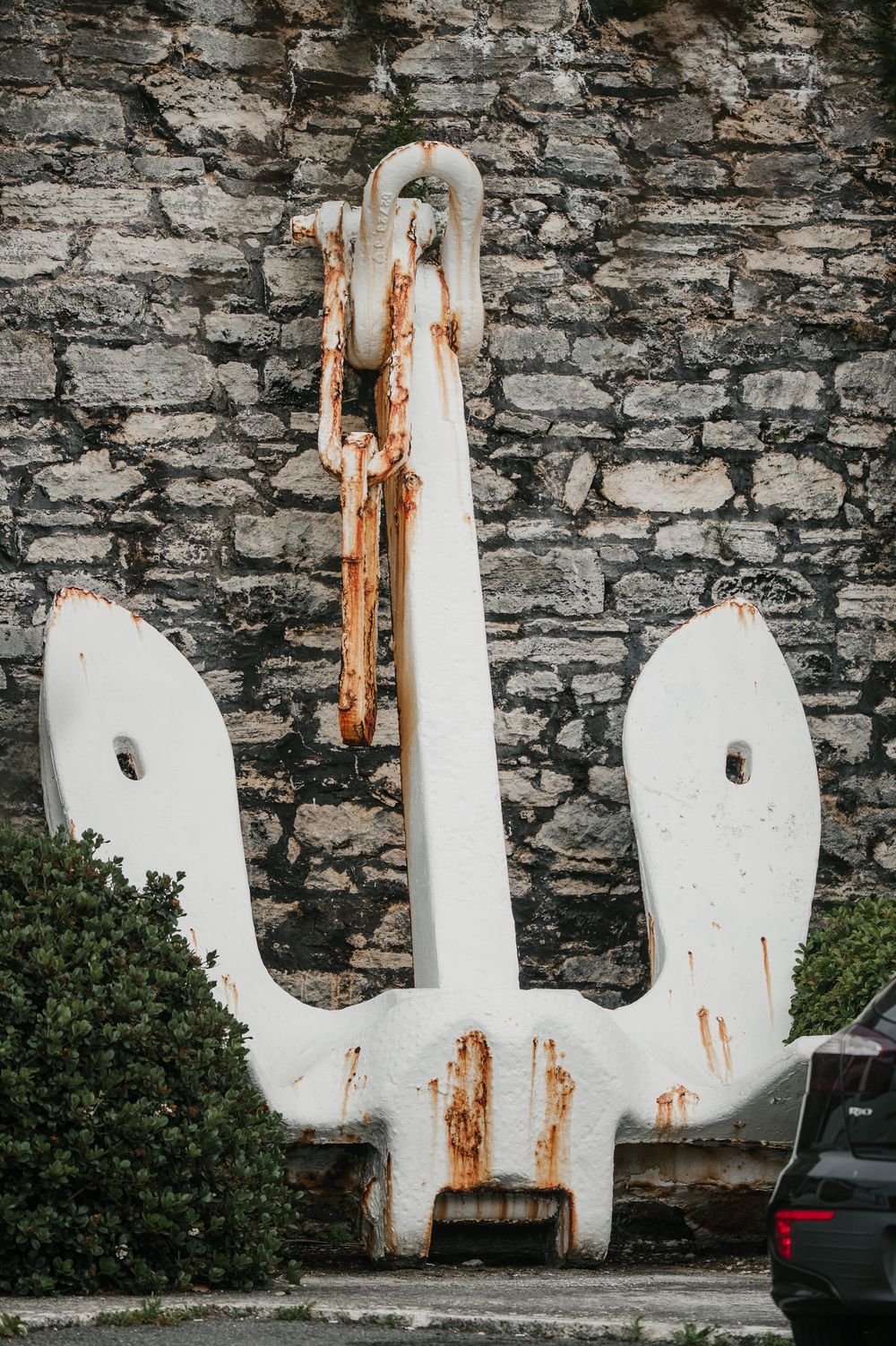 Large white rusty anchor displayed against a stone wall with a chain, serving as a nautical landmark decoration.