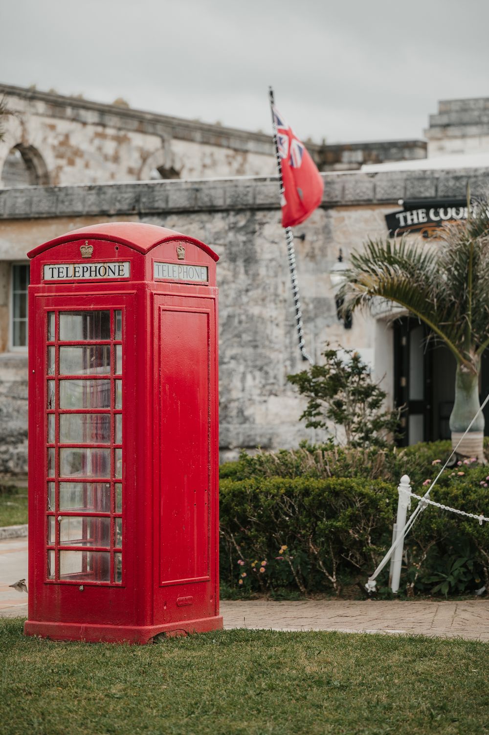 Classic red British telephone box stands on green grass beside a historic stone building with a red flag in Bermuda.
