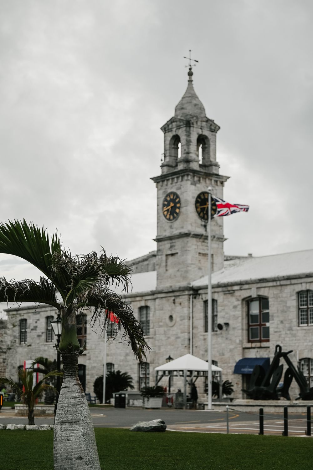 Historic clock tower building with Union Jack flag, palm trees, and anchor display under cloudy skies in Bermuda.