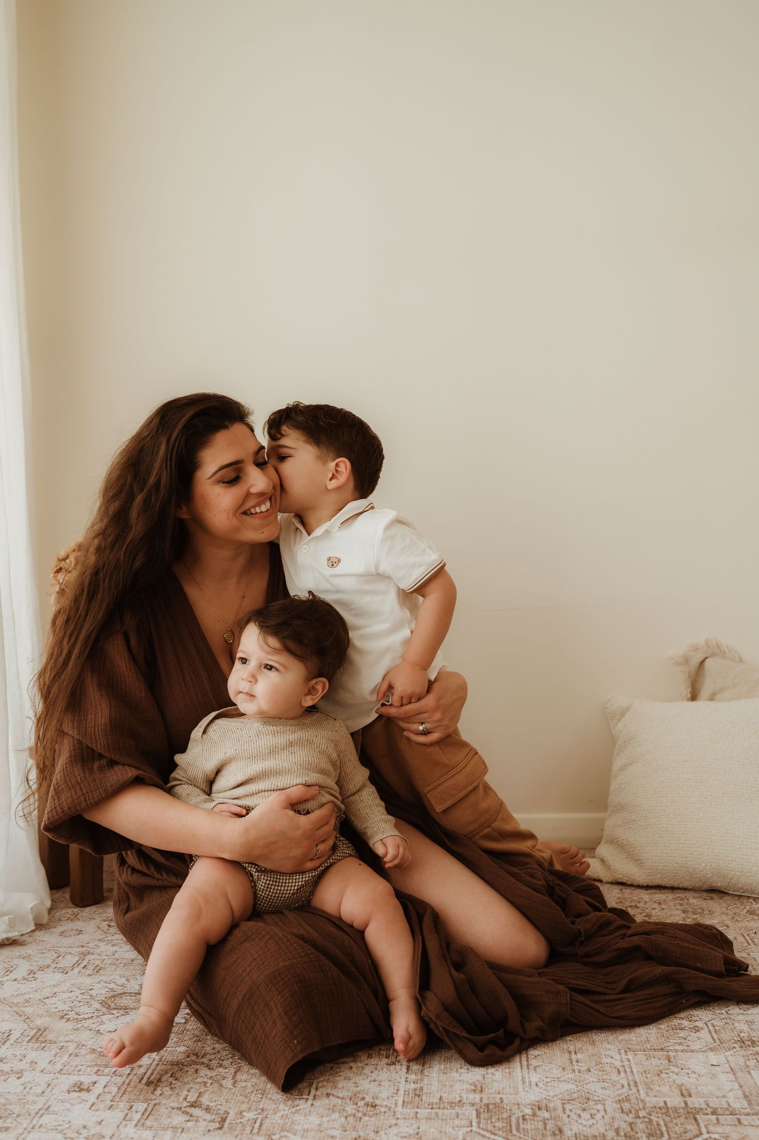 A smiling mother sits on a cream sofa with her three happy children, sharing a warm, joyful moment together.