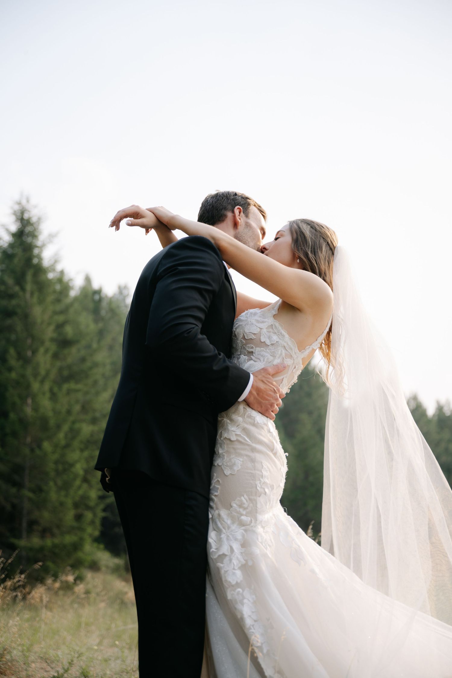 Bride and groom kissing outdoors near pine forest, veil flowing in the breeze.