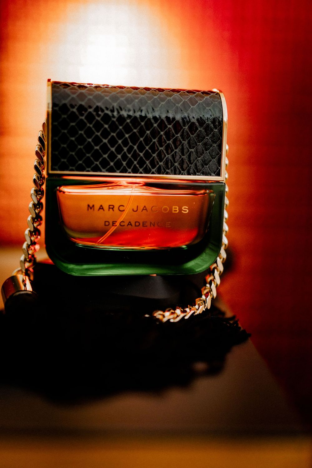 Marc Jacobs Decadence perfume bottle with black snakeskin cap and gold chain on dark background.