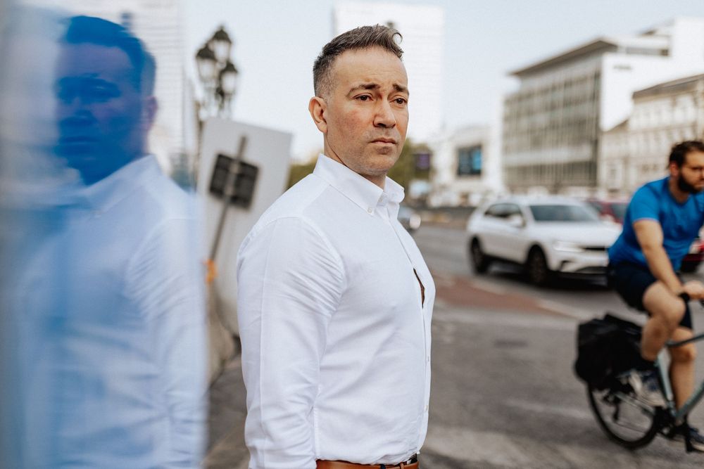A confident man in a white dress shirt stands on a city street with a bicycle rider and cars in the background.
