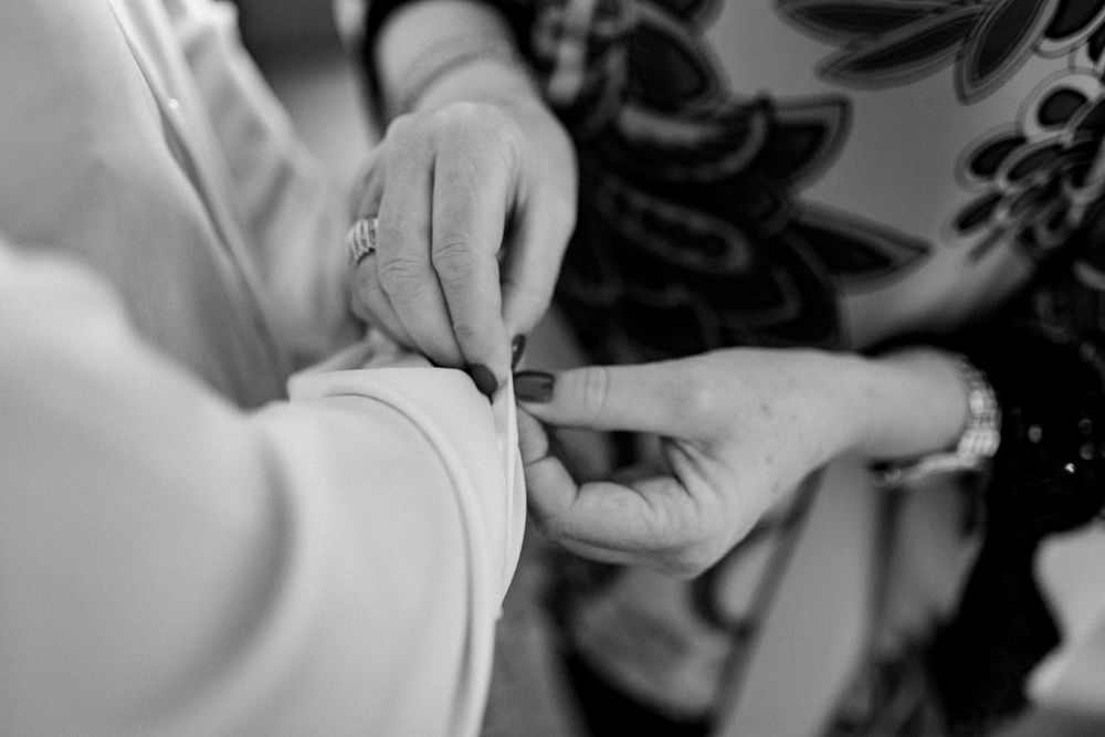 Black and white close-up of hands fastening a button on a white wedding dress sleeve.
