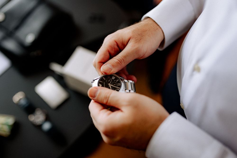 Person in white shirt adjusting a silver wristwatch with a dark dial at a desk.