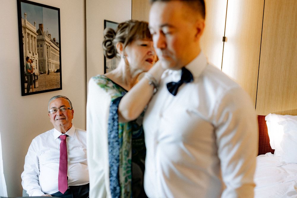 Mother adjusts grooms bow tie while father sits in background during wedding morning preparations.