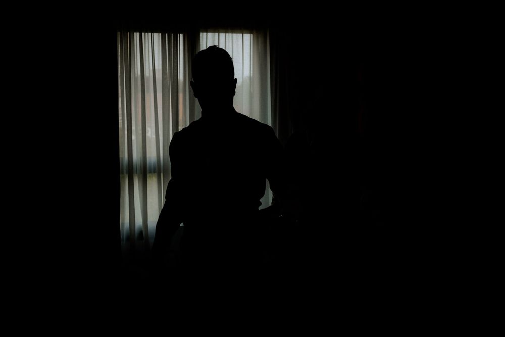 Silhouette of groom standing by curtained window in dark room before wedding ceremony.