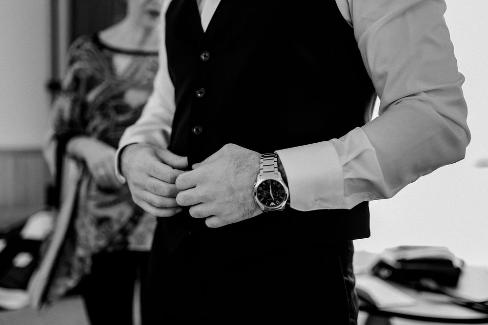 Black and white photo of a groom adjusting his black vest while wearing a luxury watch.