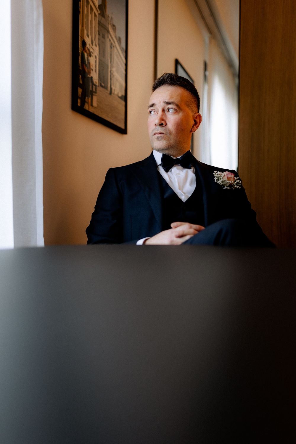 Dapper groom in navy tuxedo with bow tie and boutonniere sitting thoughtfully by window.