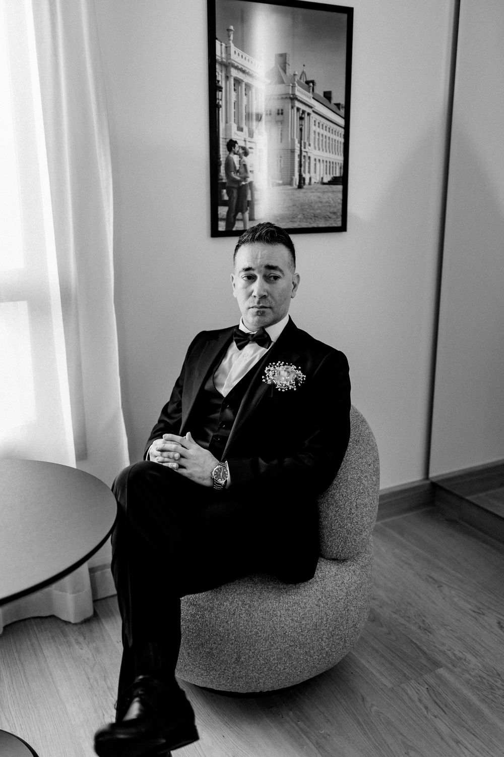 Groom in black tuxedo with bow tie seated in a chair in black and white portrait photo.