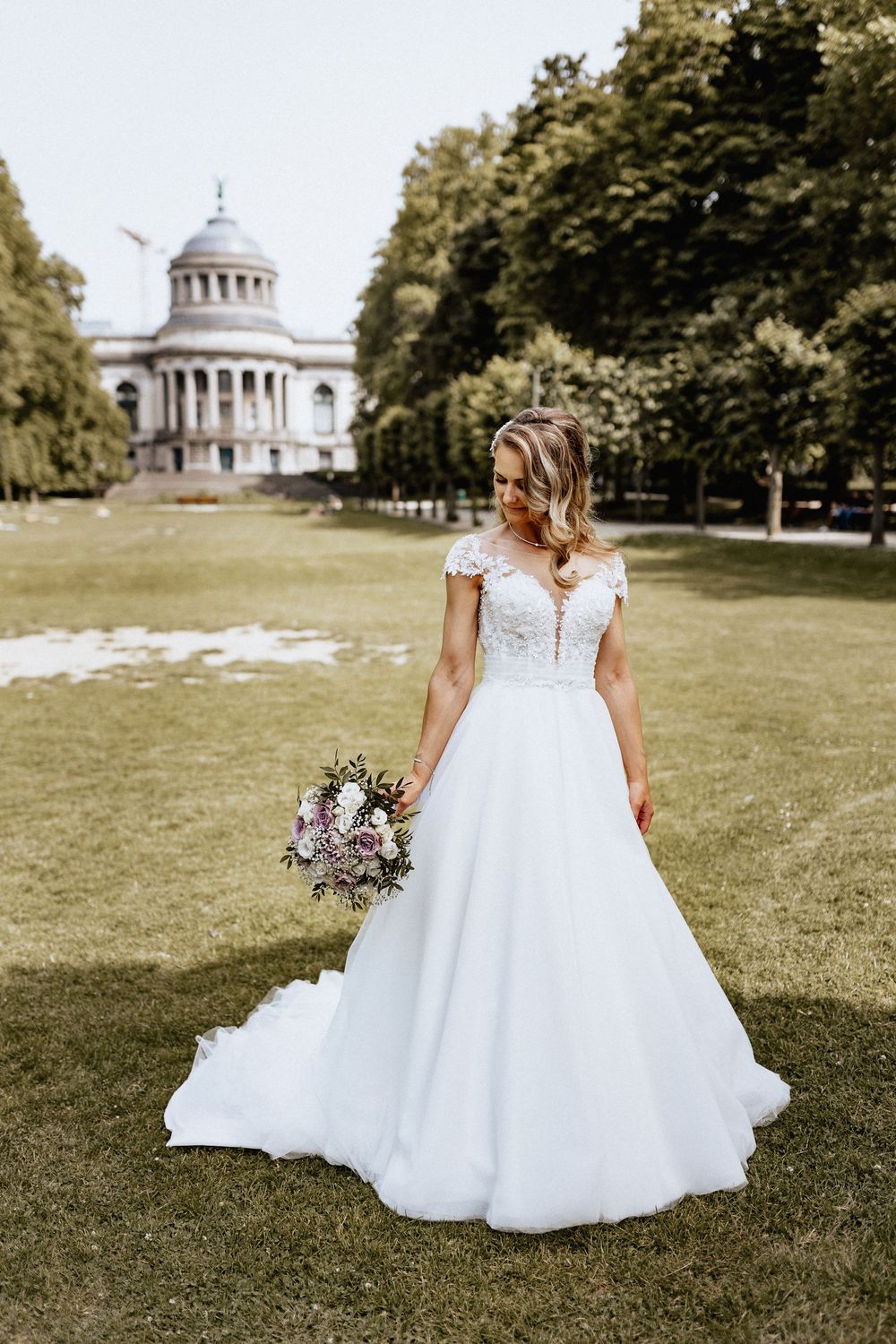 Bride in ball gown with lace bodice holds bouquet in park with domed building behind.