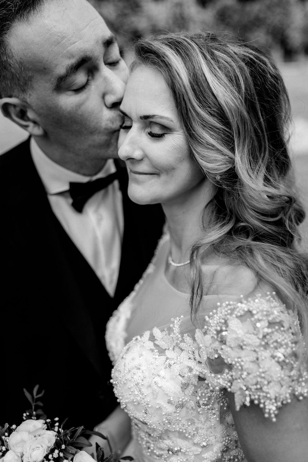 Black and white portrait of groom tenderly kissing smiling bride on cheek outdoors.