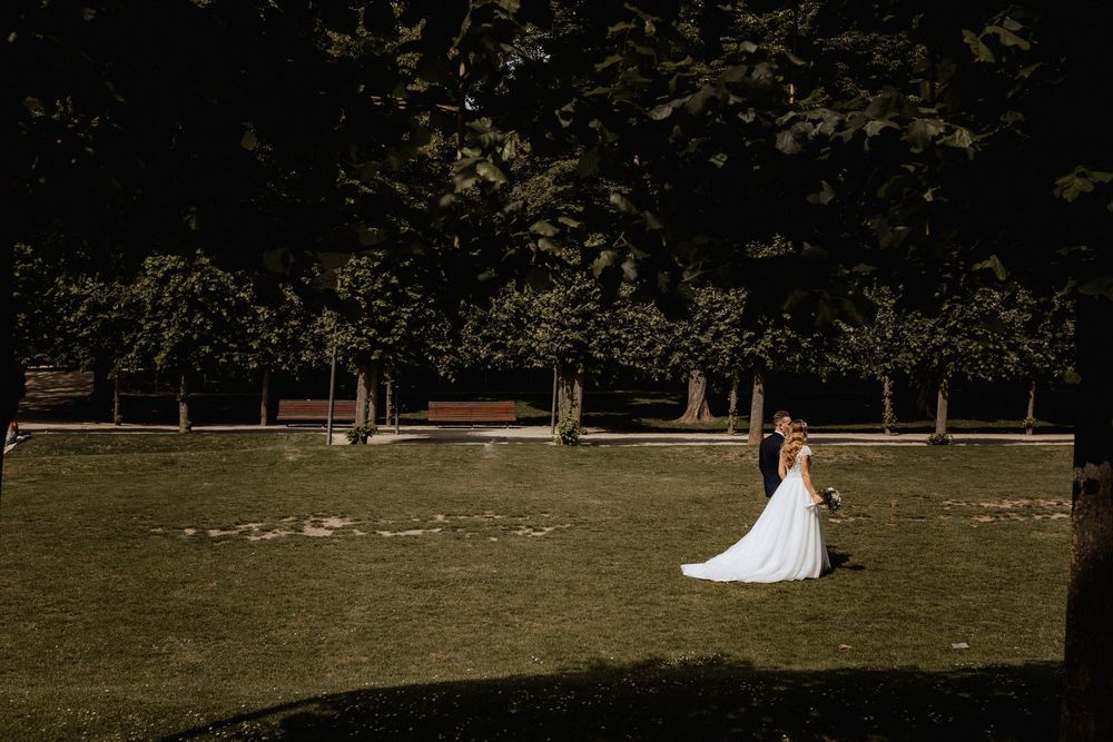 Bride and groom walk through a sunlit green park with a long flowing white wedding train.
