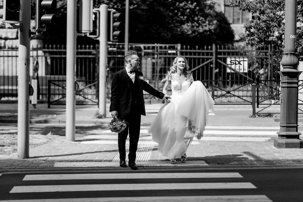 Stylish bride and groom hold hands while crossing a zebra crossing in a classic black and white urban photo.