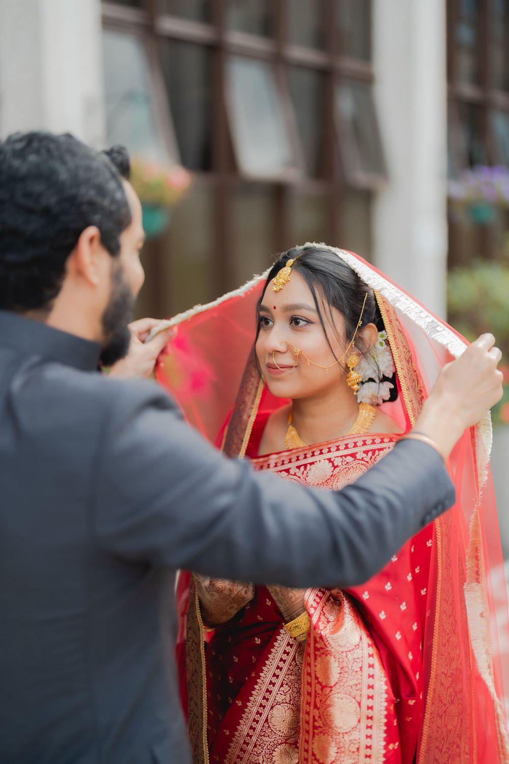 Groom lifting red dupatta veil over Indian bride in traditional red bridal lehenga with gold jewelry outdoors.
