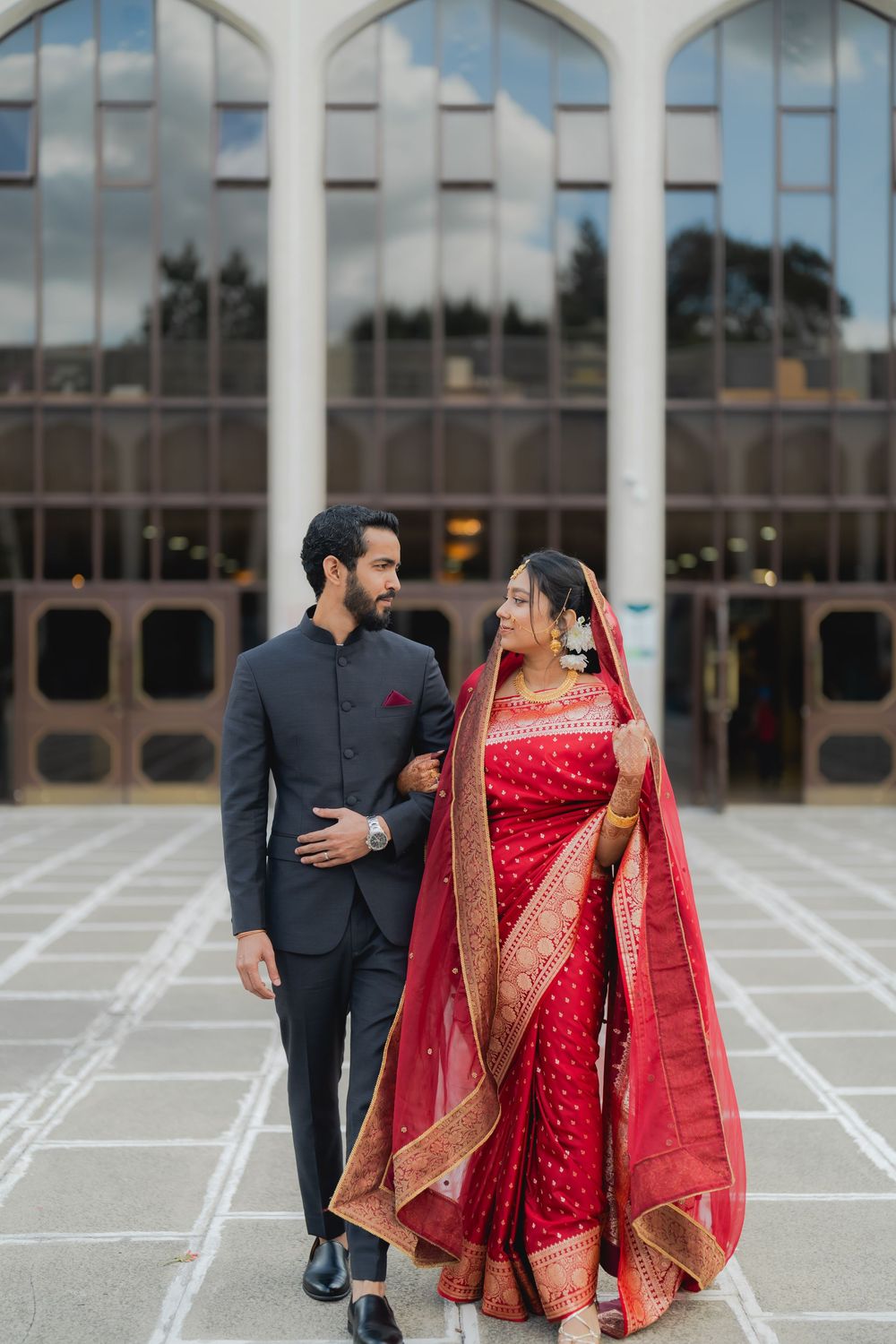 Indian bride in red saree and groom in dark suit walk together outside elegant building in wedding portrait.