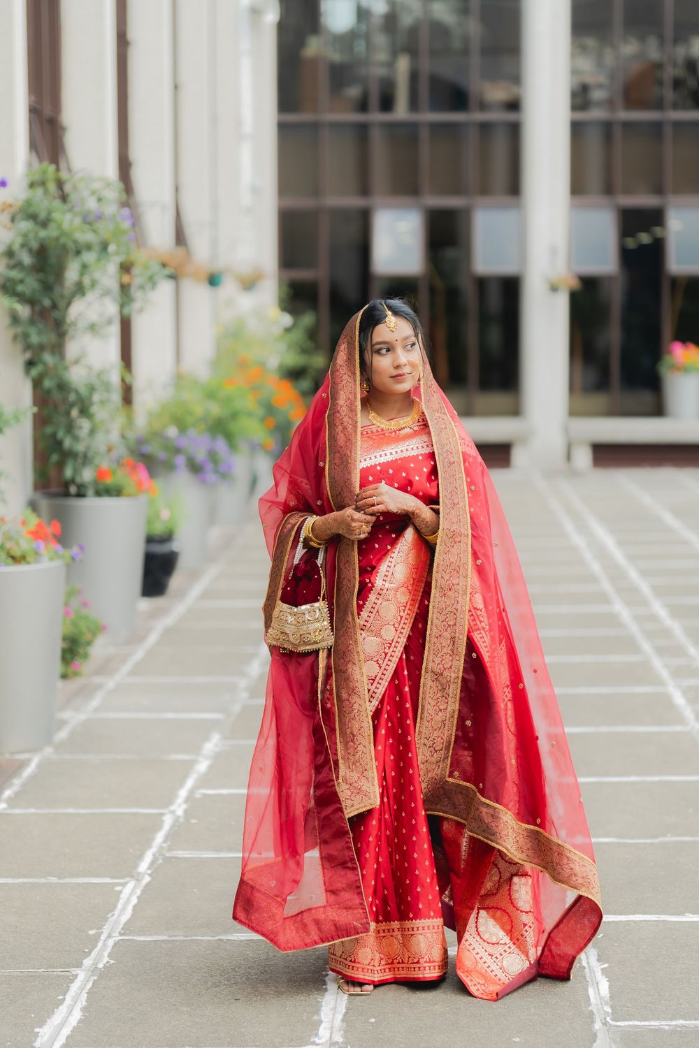 A bride in a red and gold embroidered saree with dupatta, holding a golden clutch, posing outdoors.