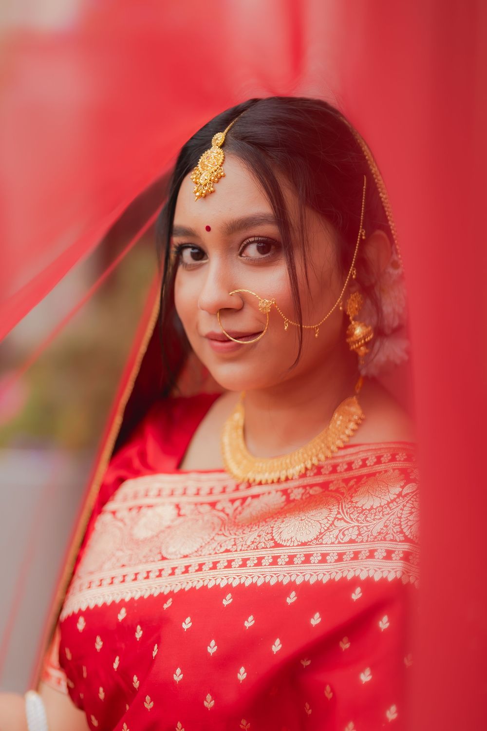 A beautiful Indian bride in a red saree with gold jewelry, bindi, and veil, posing gracefully.