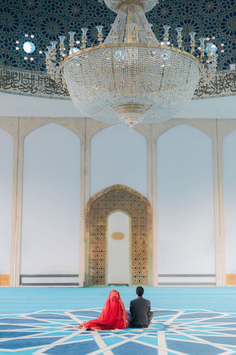 Muslim couple in red and black wedding attire sitting on blue carpet before ornate mosque mihrab under crystal chandelier.