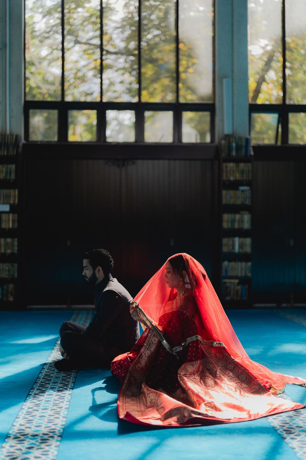 Muslim bride in red dupatta and groom in black suit sitting on blue mosque carpet with bookshelves and sunlit windows.