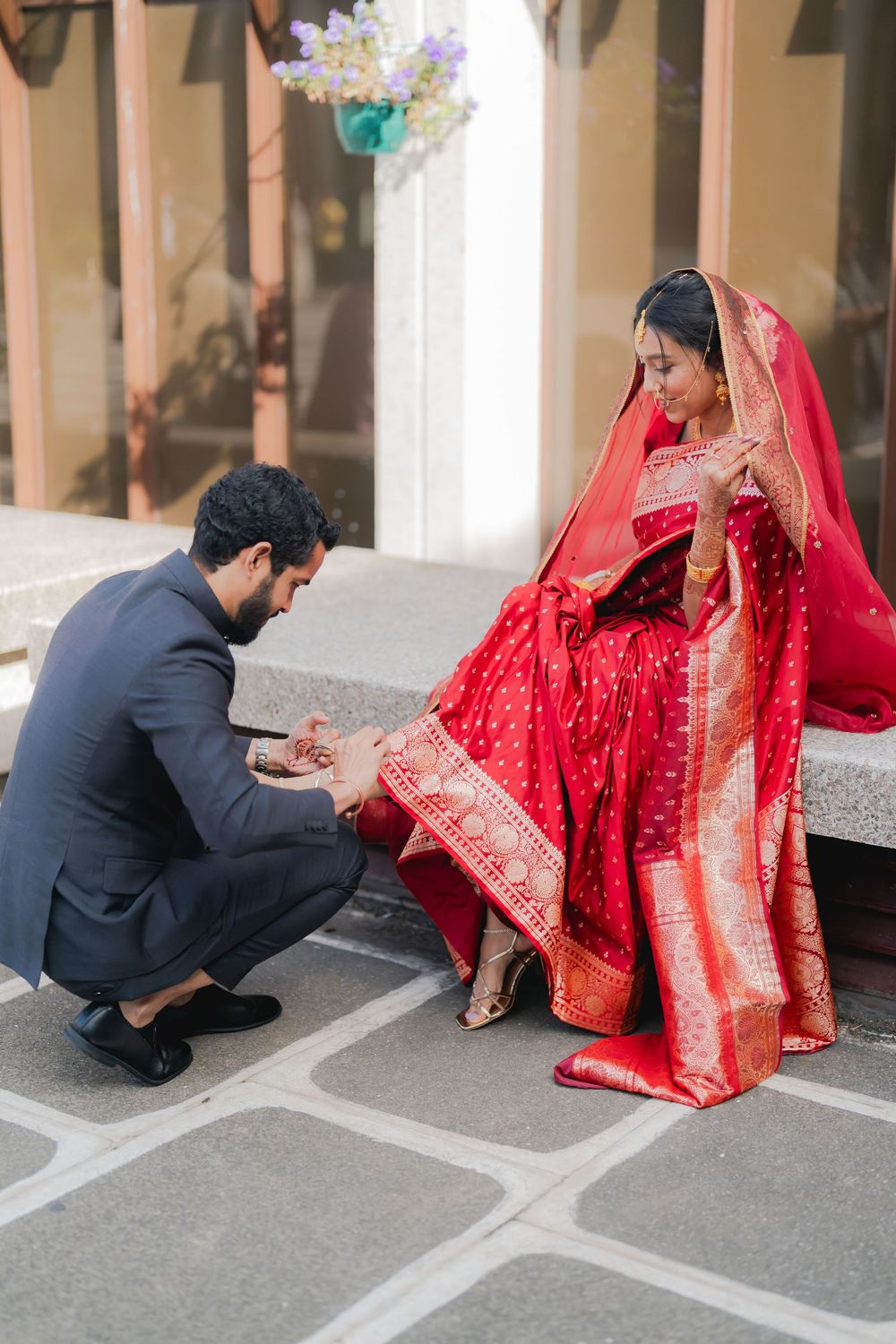 Groom in navy suit placing jewelry on bride in red silk saree during traditional Indian wedding ceremony outdoors.