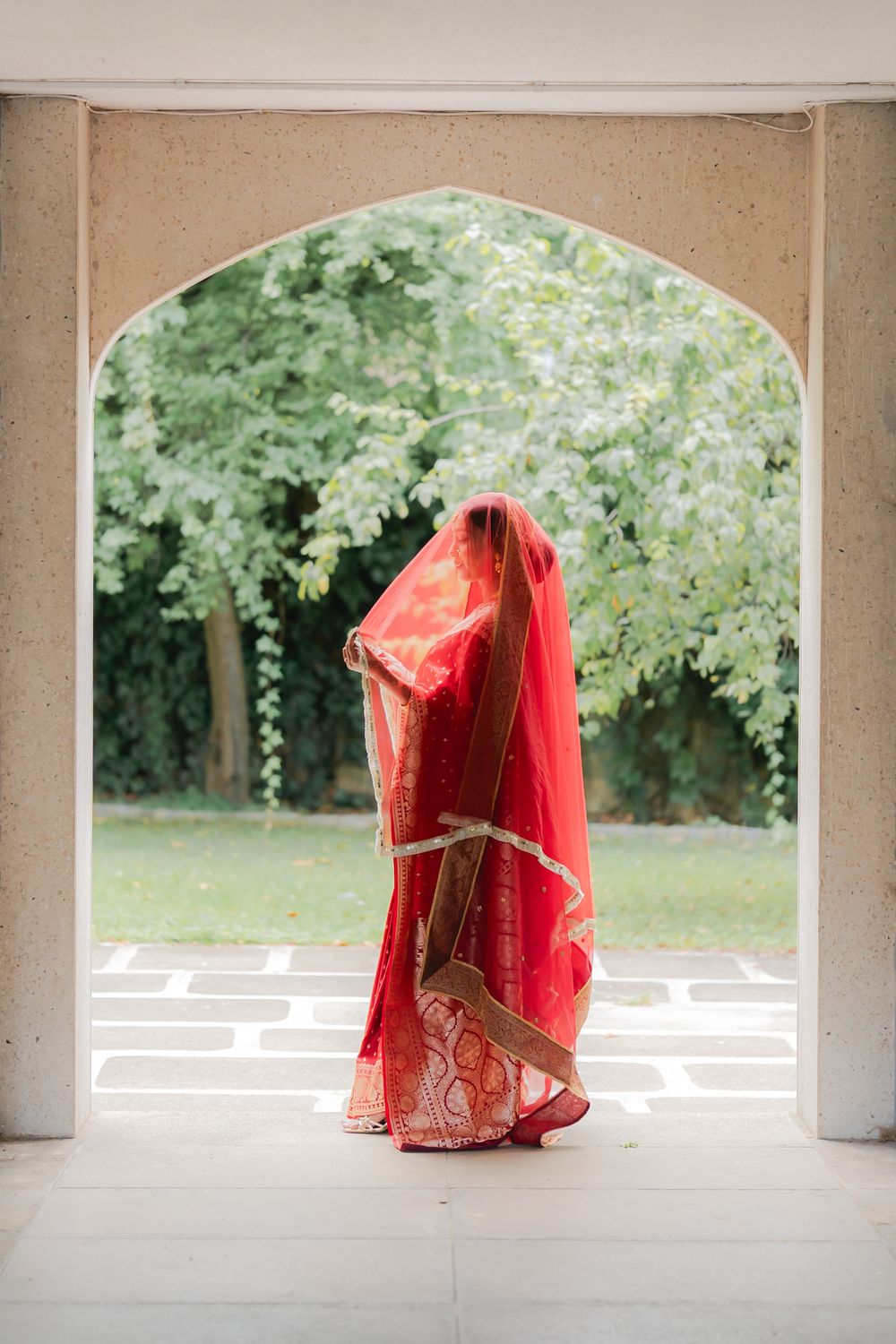 Indian bride in red saree and veil standing in ornate archway with lush green garden backdrop.