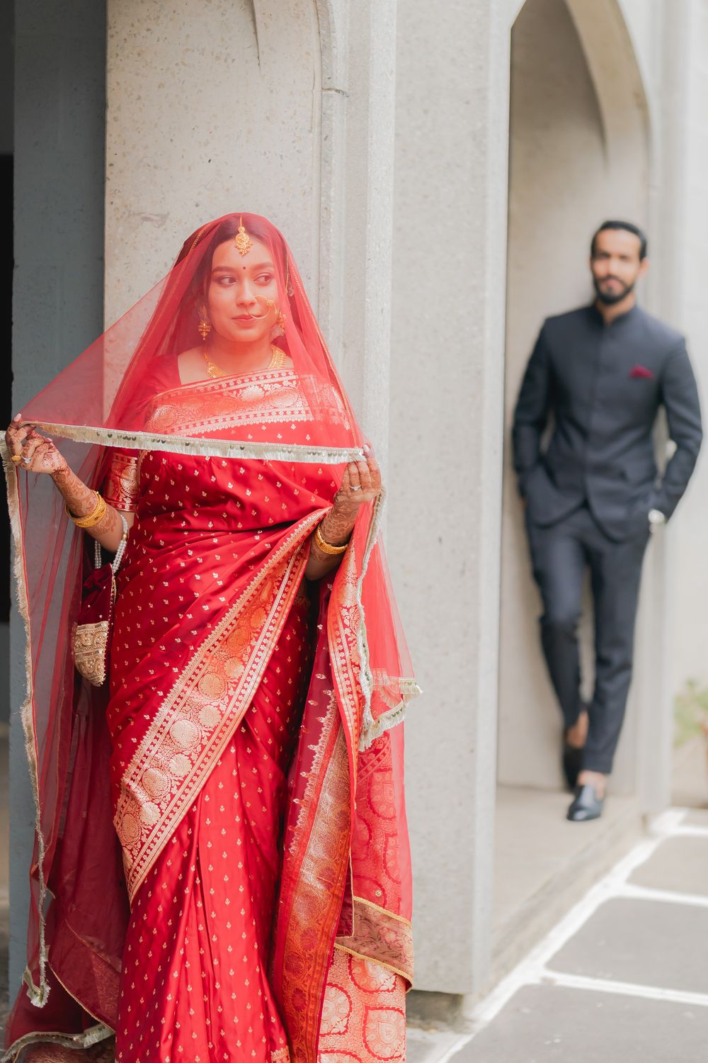 Indian bride in red bridal saree lifting veil while groom in dark suit stands in background.