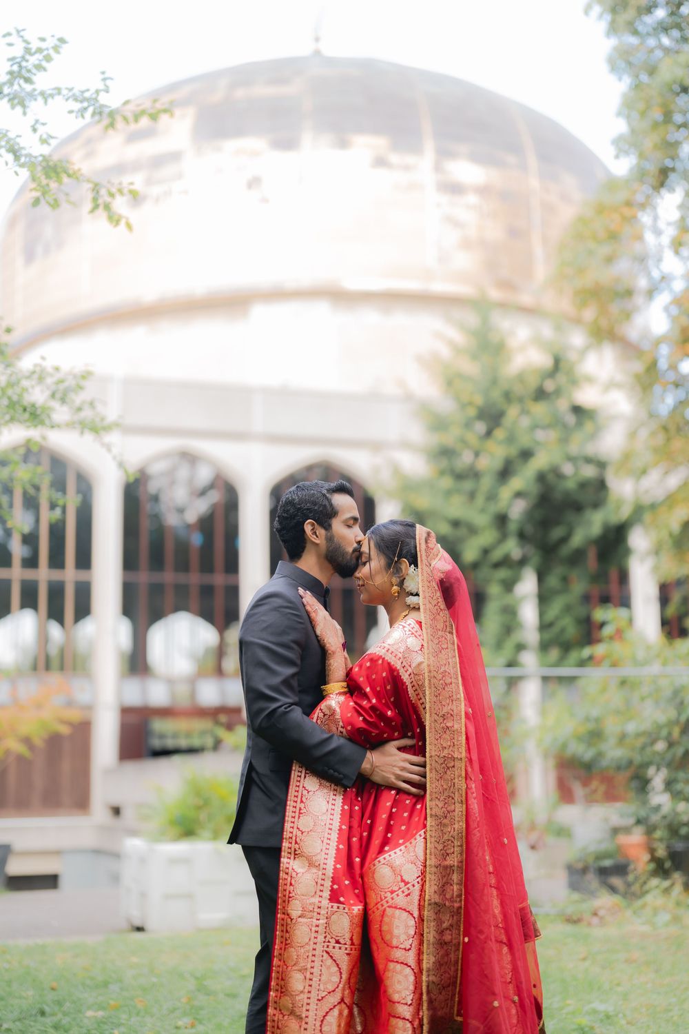 Couple sharing a kiss in traditional Indian wedding attire, bride in red saree with groom in black suit outdoors.