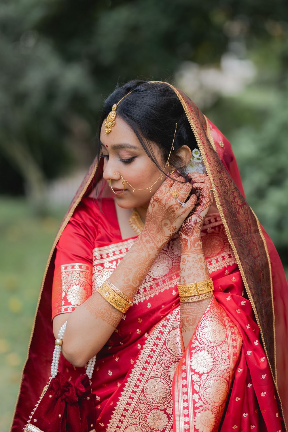 Indian bride in red Banarasi saree adjusting gold jewelry with henna-adorned hands outdoors.