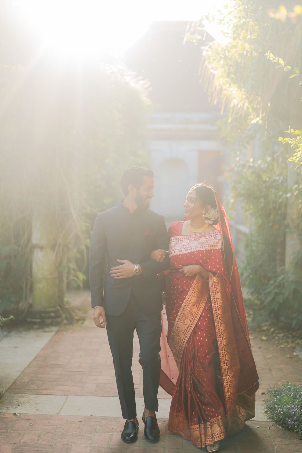 Couple in traditional Indian wedding attire walking together outdoors in soft sunlight, bride in red saree, groom in dark suit.