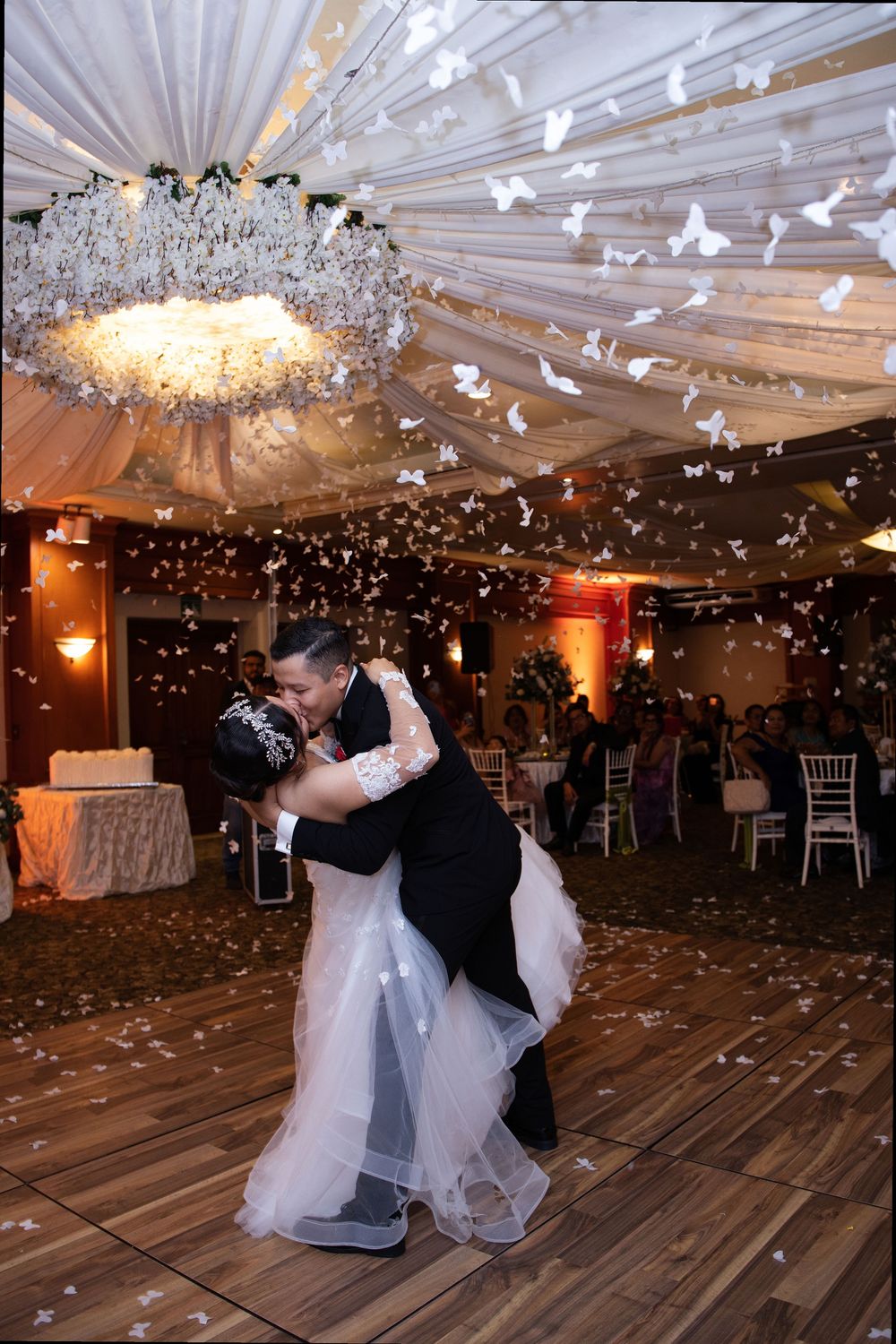 Bride and groom share romantic first dance kiss under white butterfly confetti in elegantly draped wedding reception venue.