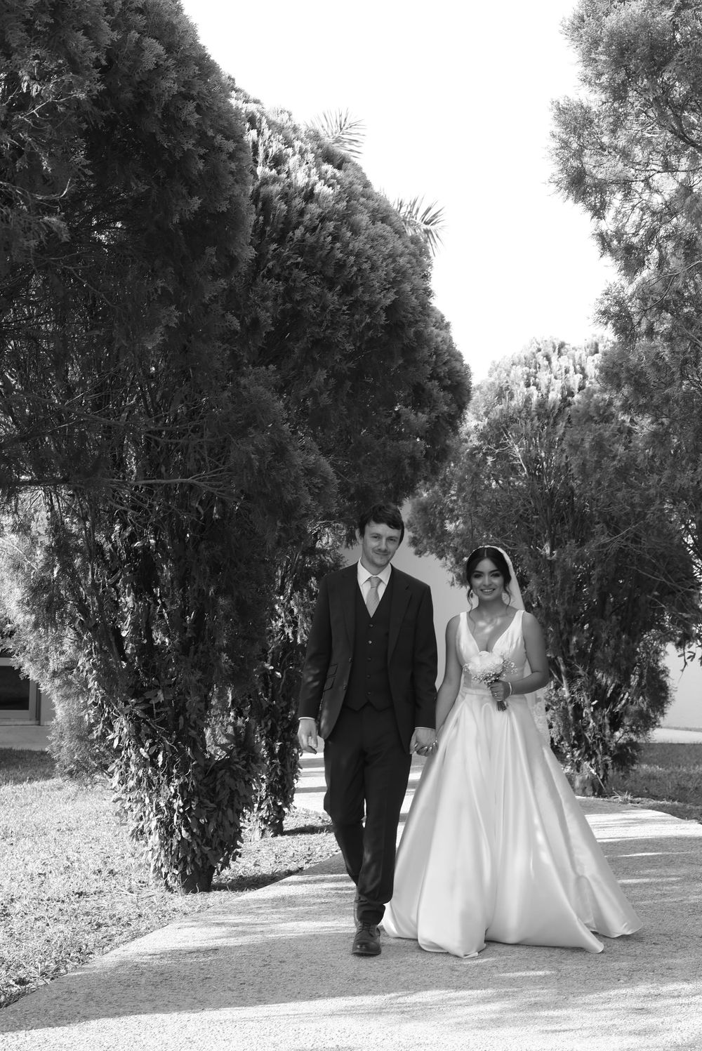 Black and white photo of a bride and groom walking hand in hand along a tree-lined path on their wedding day.