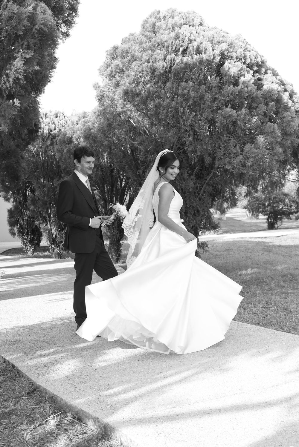 Black and white photo of a bride in flowing gown and groom in suit walking hand in hand outdoors on their wedding day.
