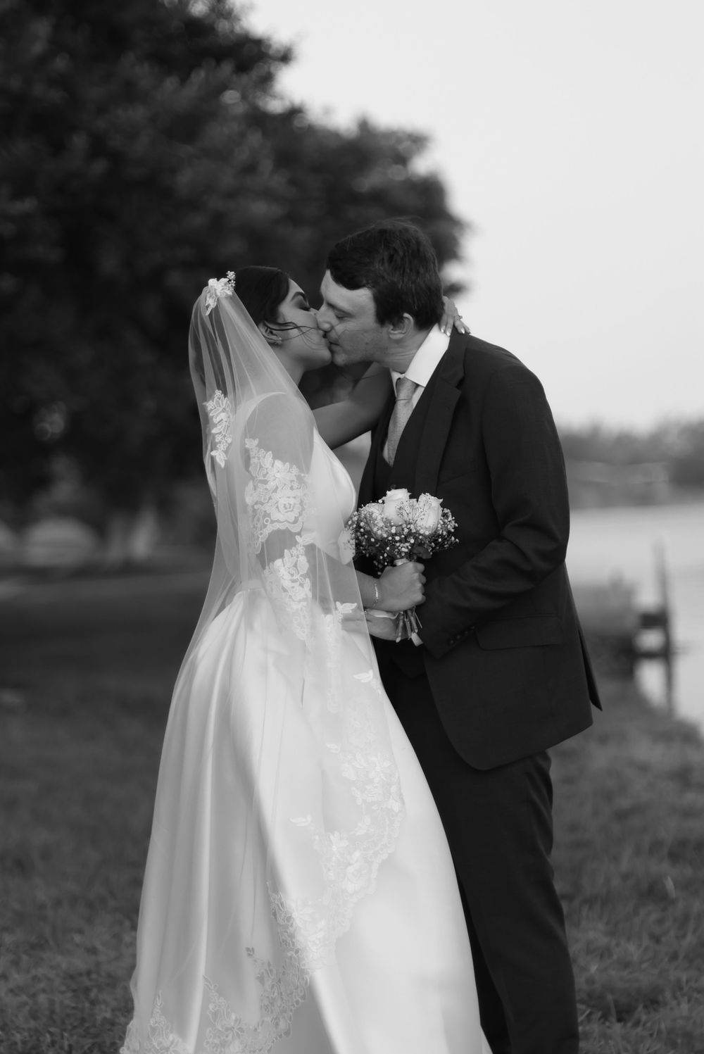 Black and white photo of a bride and groom sharing a kiss outdoors, bride holding a bouquet in a lace wedding gown.