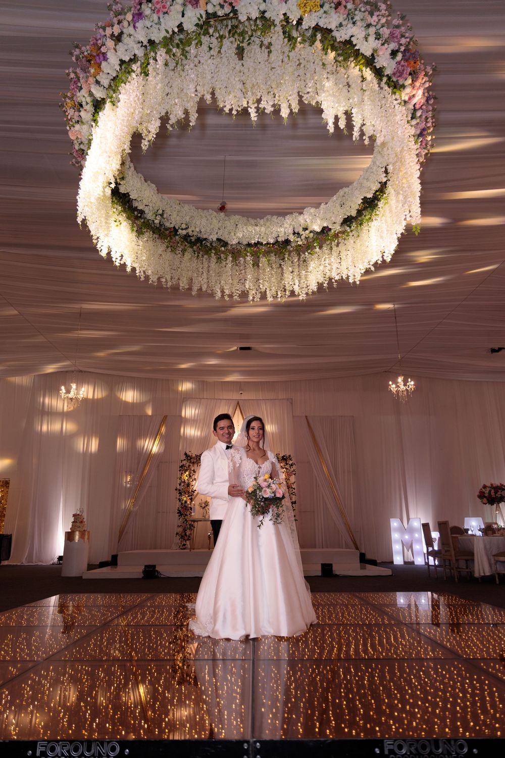 Bride and groom stand on a sparkling dance floor beneath a stunning circular floral chandelier in an elegant wedding venue.